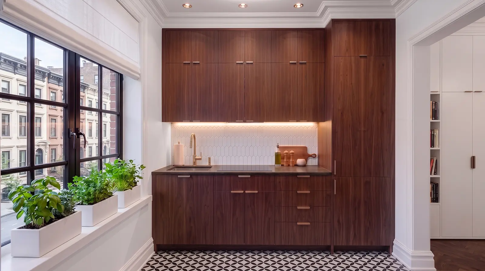 Manhattan studio apartment kitchenette with walnut cabinetry, compact appliances, stone counters, mosaic floor and geometric white ceramic tile wall.